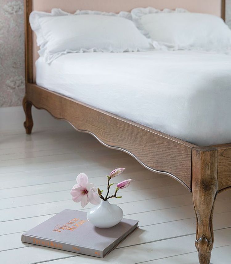 Wooden bed with white bedding, next to a small vase with pink flowers and a closed book on a white wooden floor.