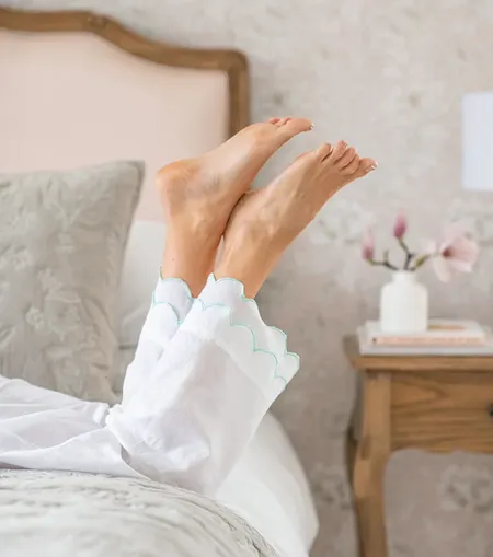 Feet in white pajama pants with scalloped edges are playfully raised in the air, resting on a bed with a soft, neutral-toned bedroom background.