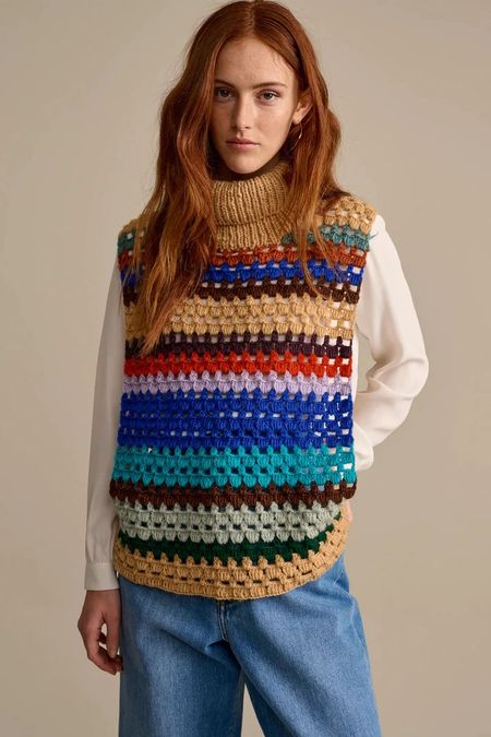 Person wearing a multicolored crocheted vest over a white shirt and blue jeans, standing against a plain background.
