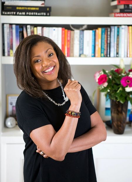 Smiling woman in a black dress stands in front of a bookshelf with colorful books and flowers, wearing a beaded bracelet and necklace.