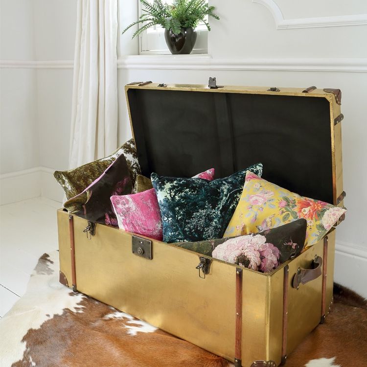 Open gold trunk filled with colorful velvet pillows, featuring floral and abstract patterns, placed on a cowhide rug in a bright room.