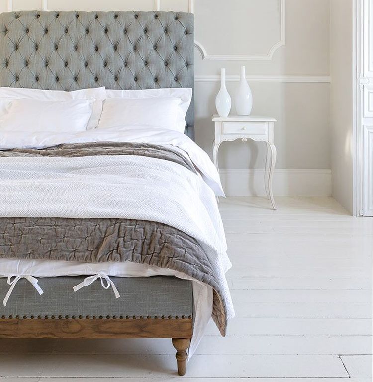 Elegant bedroom with a tufted headboard, white bedding, and a gray blanket. A small white side table holds two decorative vases.