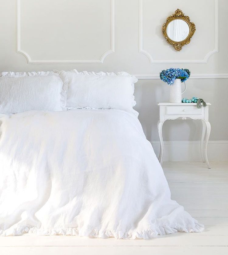 Elegant bedroom with a white ruffled bedspread, ornate mirror, and a side table holding a vase of blue flowers against a paneled wall.