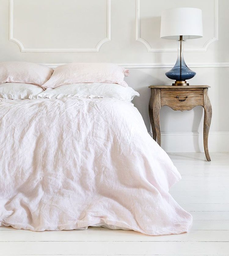 A cozy bedroom with a neatly made bed, light pink bedding, a wooden side table, and a blue lamp, all against a white paneled wall.