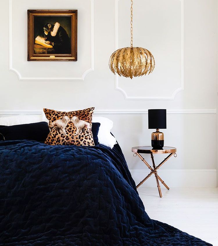 Elegant bedroom with a navy quilt, leopard print pillow, gold leaf chandelier, black lamp on a round table, and a painting on the wall.