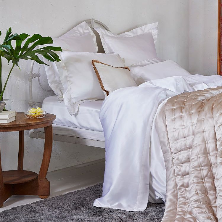 A cozy bedroom with a neatly made bed featuring white pillows and sheets, a beige quilt, a wooden side table, and a green plant.