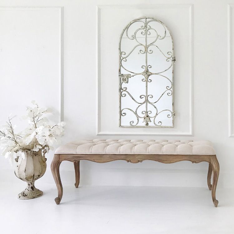 Elegant room with a tufted bench, ornate arched mirror, and decorative vase with white flowers against a white paneled wall.