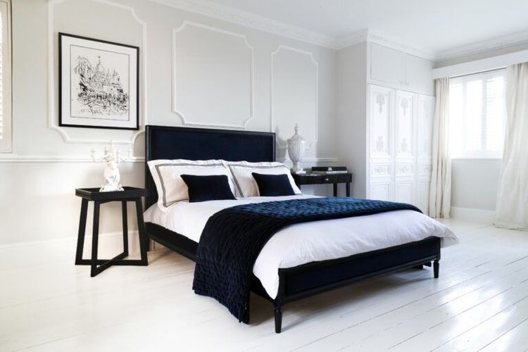 Modern bedroom with a black bed, white bedding, navy accents, and elegant decor. Light gray walls, white floor, and framed artwork above the bed.