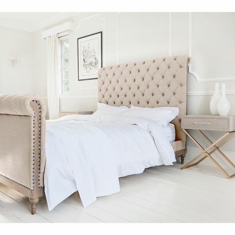 Elegant bedroom with a tufted beige bed, white bedding, and a gold-accented nightstand. Bright natural light from a window.