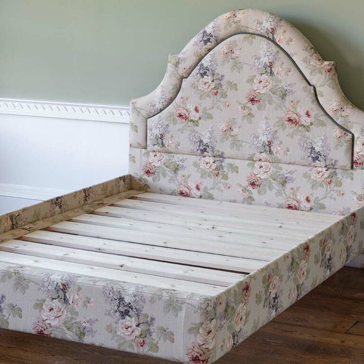 Floral-patterned bed frame with an ornate headboard, featuring a light wood slatted base, set against a pale green wall with white molding.