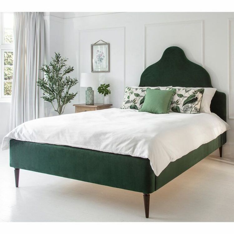 Elegant bedroom with a green upholstered bed, white bedding, decorative pillows, a potted plant, and a wooden side table with a lamp.