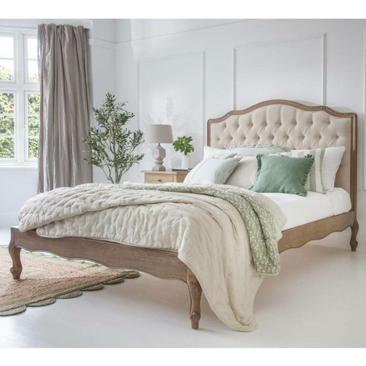 Elegant bedroom with a tufted bed, green and white pillows, light quilt, wooden nightstand, lamp, plant, and large window with curtains.