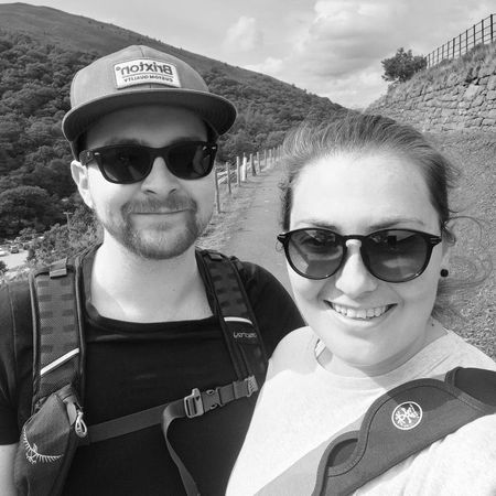 A smiling couple wearing sunglasses and backpacks take a selfie on a sunny day, with a hillside and trees in the background.