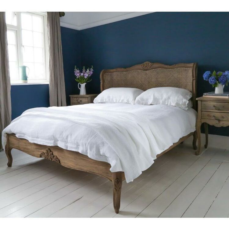 Elegant bedroom with a wooden bed, white bedding, and blue walls. Two nightstands hold vases of purple flowers by a window with beige curtains.