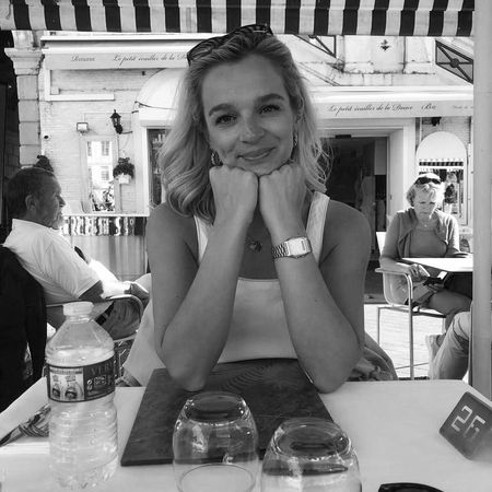 Woman smiling, resting her chin on her hands at an outdoor café table. Bottled water and glasses are on the table. Black and white photo.
