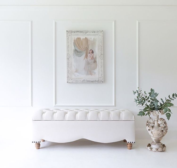Elegant white room with a tufted bench, framed artwork above, and a decorative vase with greenery on the right.
