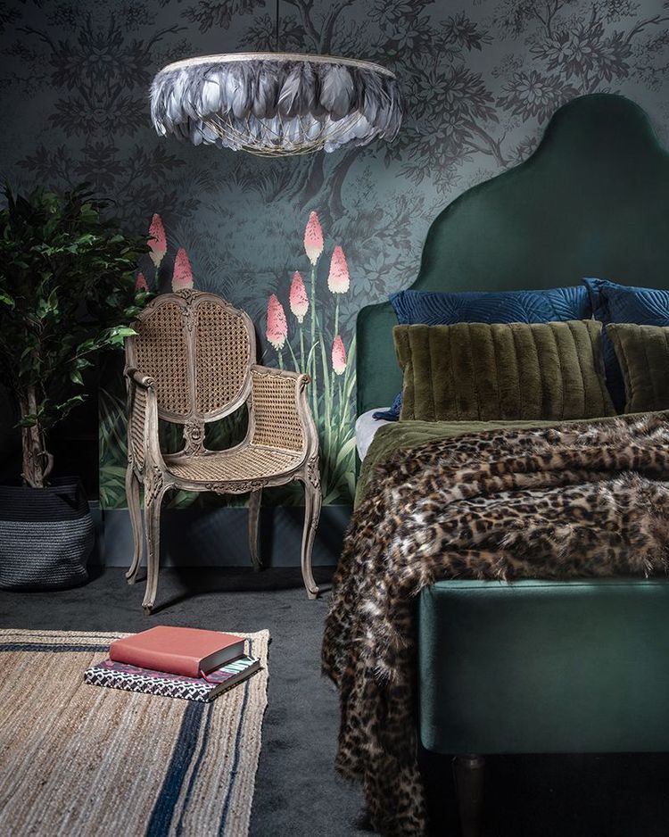 Elegant bedroom with a green velvet bed, floral wallpaper, feathered chandelier, ornate chair, and a leopard print throw. Books lie on the rug.