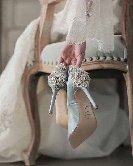 A bride in a lace gown holds elegant blue high heels adorned with intricate crystal embellishments while sitting on a vintage chair.