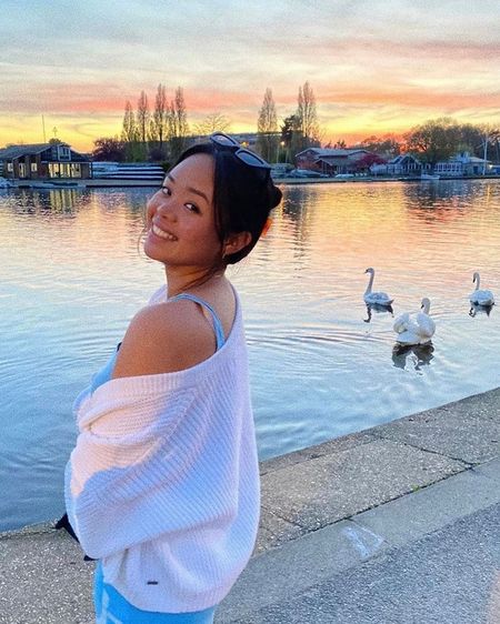 Woman smiling by a lake during sunset, wearing a white sweater, with swans in the water and colorful sky in the background.