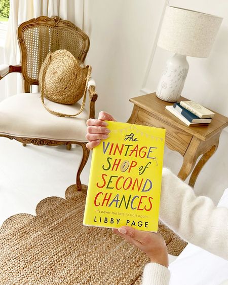 Person holding "The Vintage Shop of Second Chances" book in a cozy room with a chair, woven bag, and a wooden side table with a lamp.
