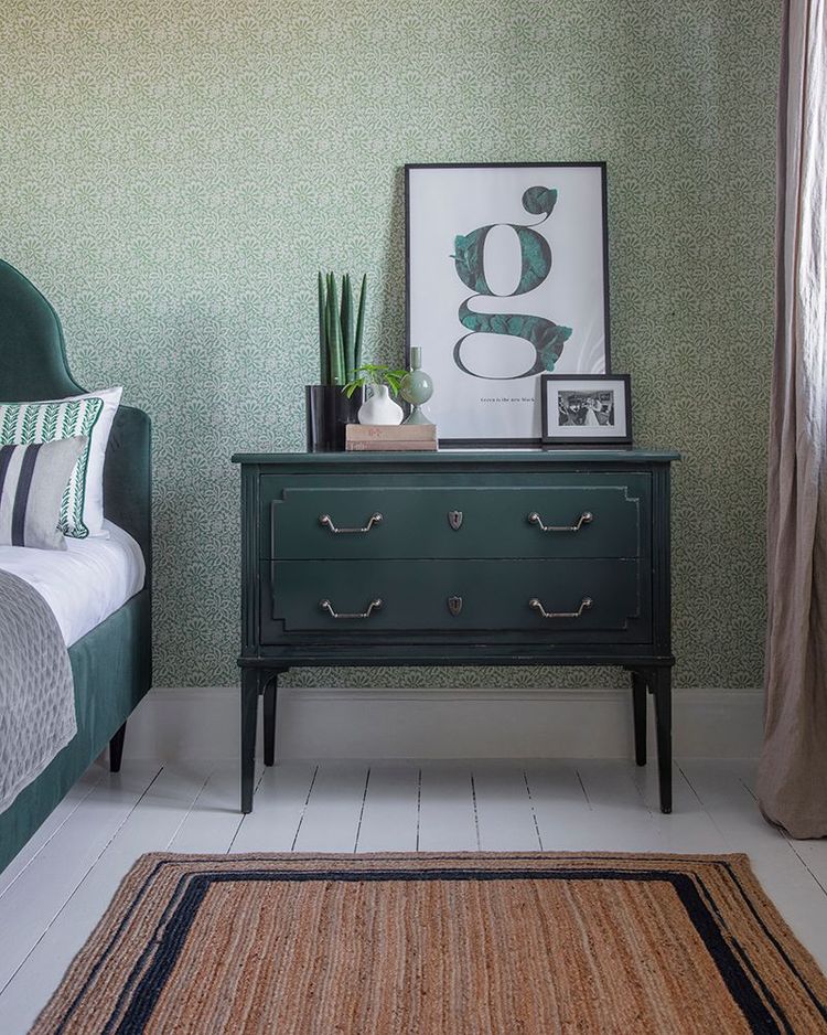 Bedroom with a green nightstand, framed art, potted plants, and a bed with patterned pillows. The room has patterned wallpaper and a striped rug.