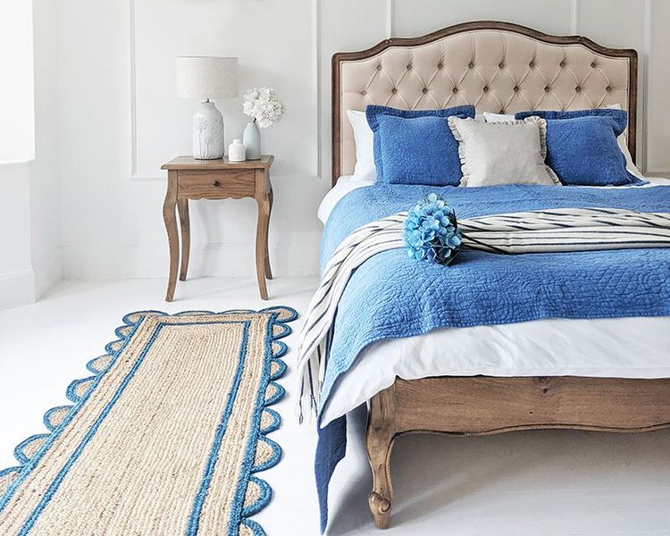 Elegant bedroom with a wooden bed, blue bedding, and a matching runner. A side table holds a lamp and vase with flowers. White walls enhance brightness.