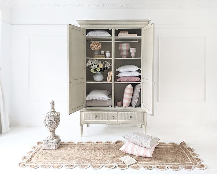 Open cabinet with neatly arranged pillows and decor items, next to a decorative urn on a rug in a white room.