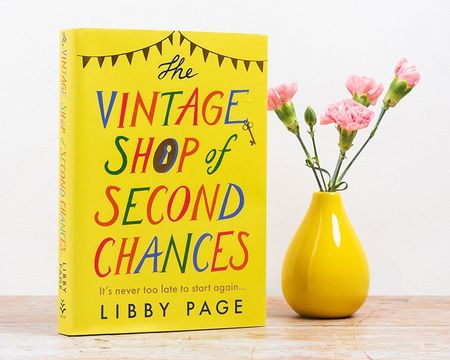 Yellow book titled "The Vintage Shop of Second Chances" by Libby Page next to a yellow vase with pink flowers on a wooden surface.