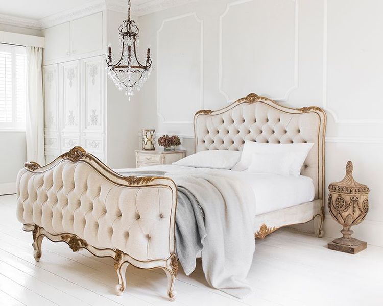 Elegant bedroom with a tufted beige bed, ornate wood details, a crystal chandelier, and a decorative urn on white wooden floors.