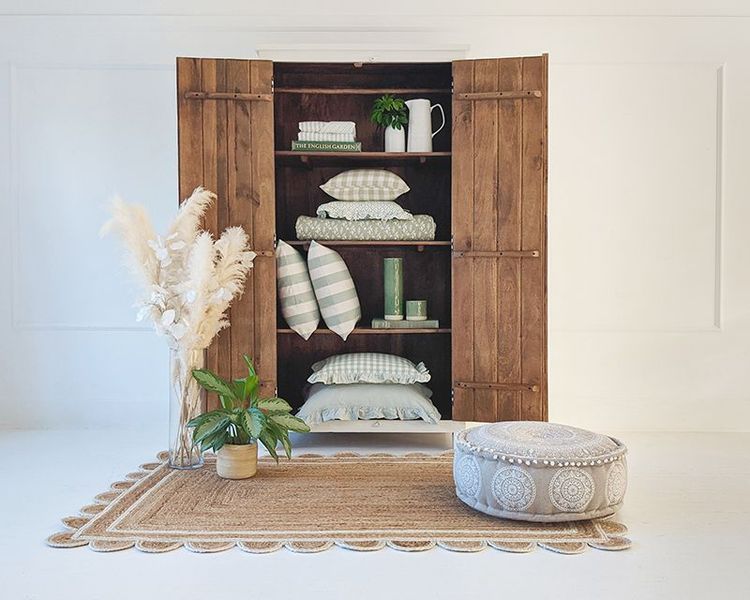 Wooden cabinet with open doors displaying pillows, books, and a white pitcher. Nearby, a pouf, plants, and a textured rug enhance the decor.