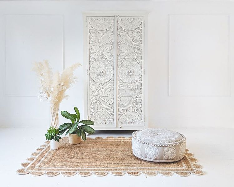 Minimalist room with a white ornate cabinet, pampas grass in a vase, green plants, a round patterned pouf, and a textured rug on the floor.