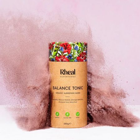 A cylindrical container of Rheal Superfoods Balance Tonic with colorful floral design, surrounded by a burst of powder on a pink background.