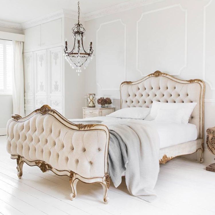 Elegant bedroom with a vintage-style tufted bed, ornate chandelier, and soft gray blanket. White walls and furniture create a serene atmosphere.