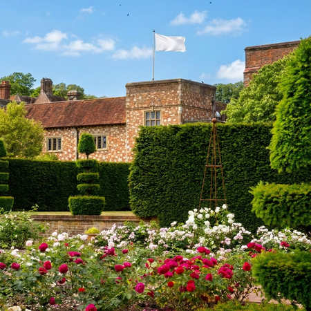 Historic brick building with a white flag, surrounded by lush gardens and colorful flowers under a clear blue sky.
