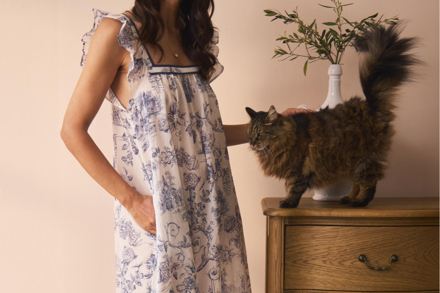 A woman wearing a blue and white toile print pyjama dress with frill shoulders, with her hand in the pocket while stroking a cat.