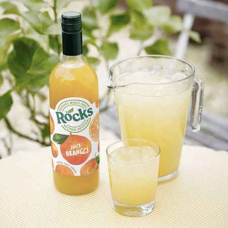 A bottle of Rocks Juicy Oranges juice next to a pitcher and glass filled with orange juice on a checkered tablecloth. Leaves in the background.