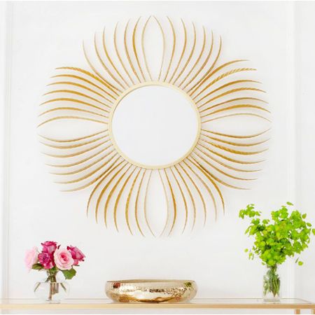 Gold sunburst mirror on a white wall above a shelf with pink roses, greenery, and a gold bowl.