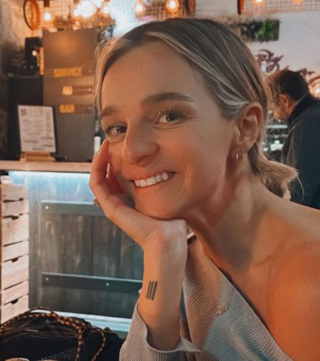 Smiling woman with a tattoo of three vertical lines on her forearm, sitting in a cozy, warmly lit restaurant.
