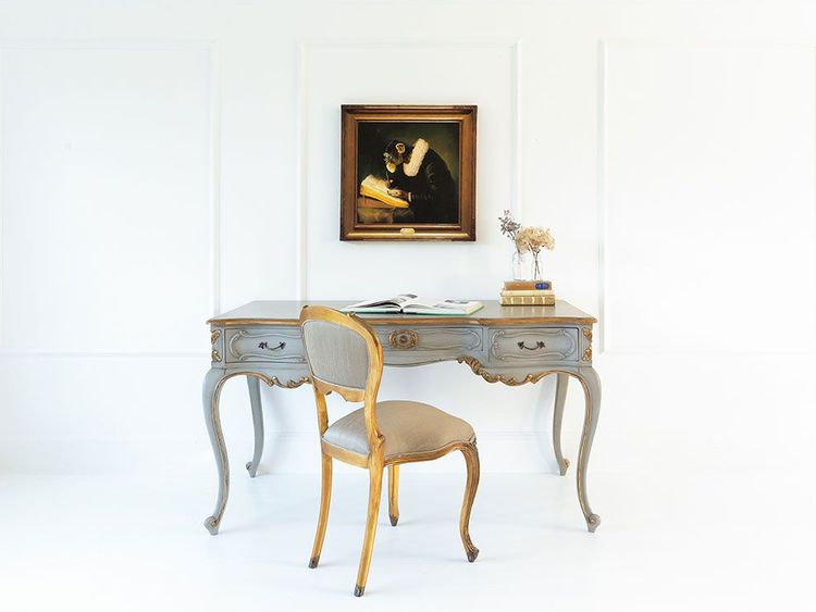 Elegant vintage desk with cabriole legs, a cushioned chair, and a framed painting of a monkey reading, set against a white wall.