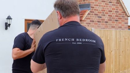 Two men in black shirts carry a large cardboard box labeled "French Bedroom Est. 2006" in a residential area.