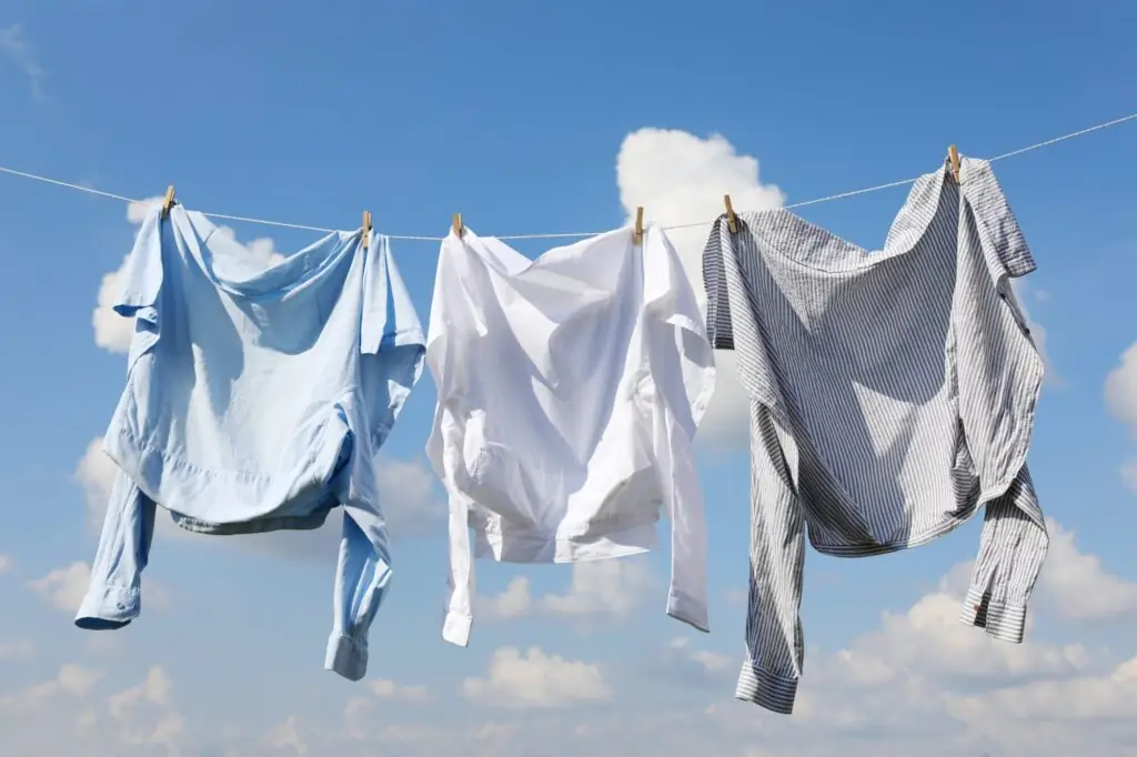 Three shirts, blue, white, and striped, hang on a clothesline against a clear blue sky with scattered clouds.