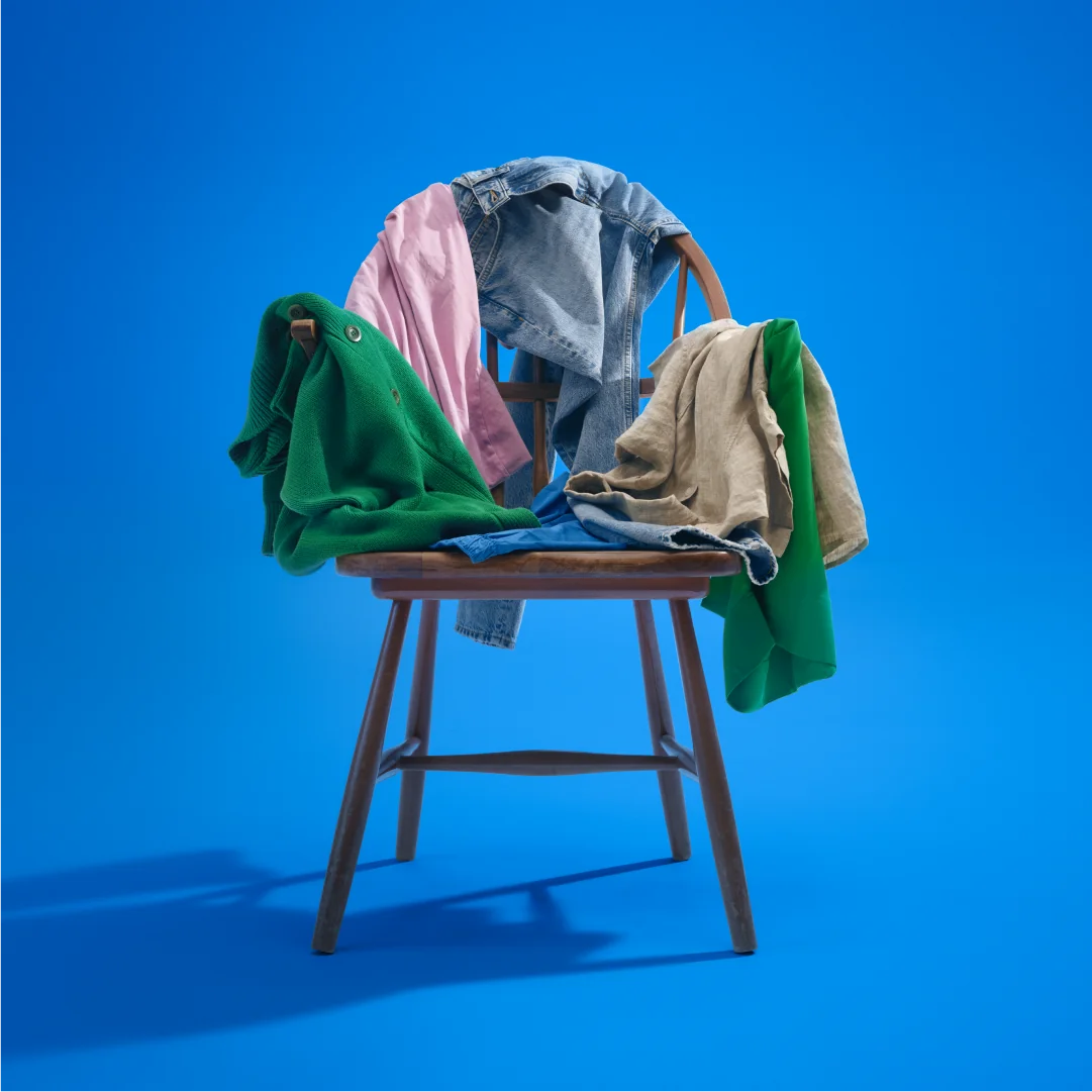 A wooden chair with various colorful clothes, including a green sweater and denim jacket, draped over it against a bright blue background.