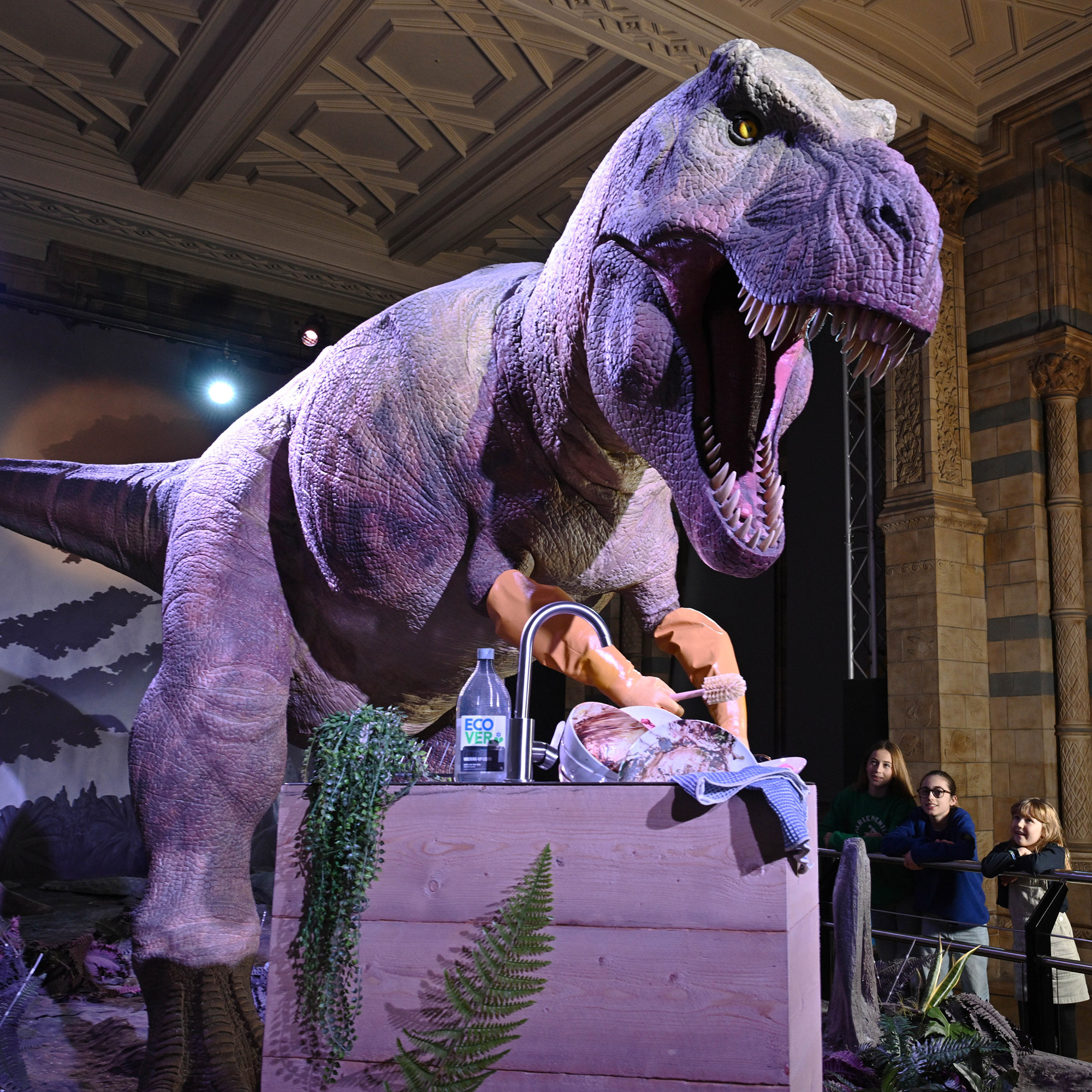 A Tiranosaurus wearing gloves while doing the dishes