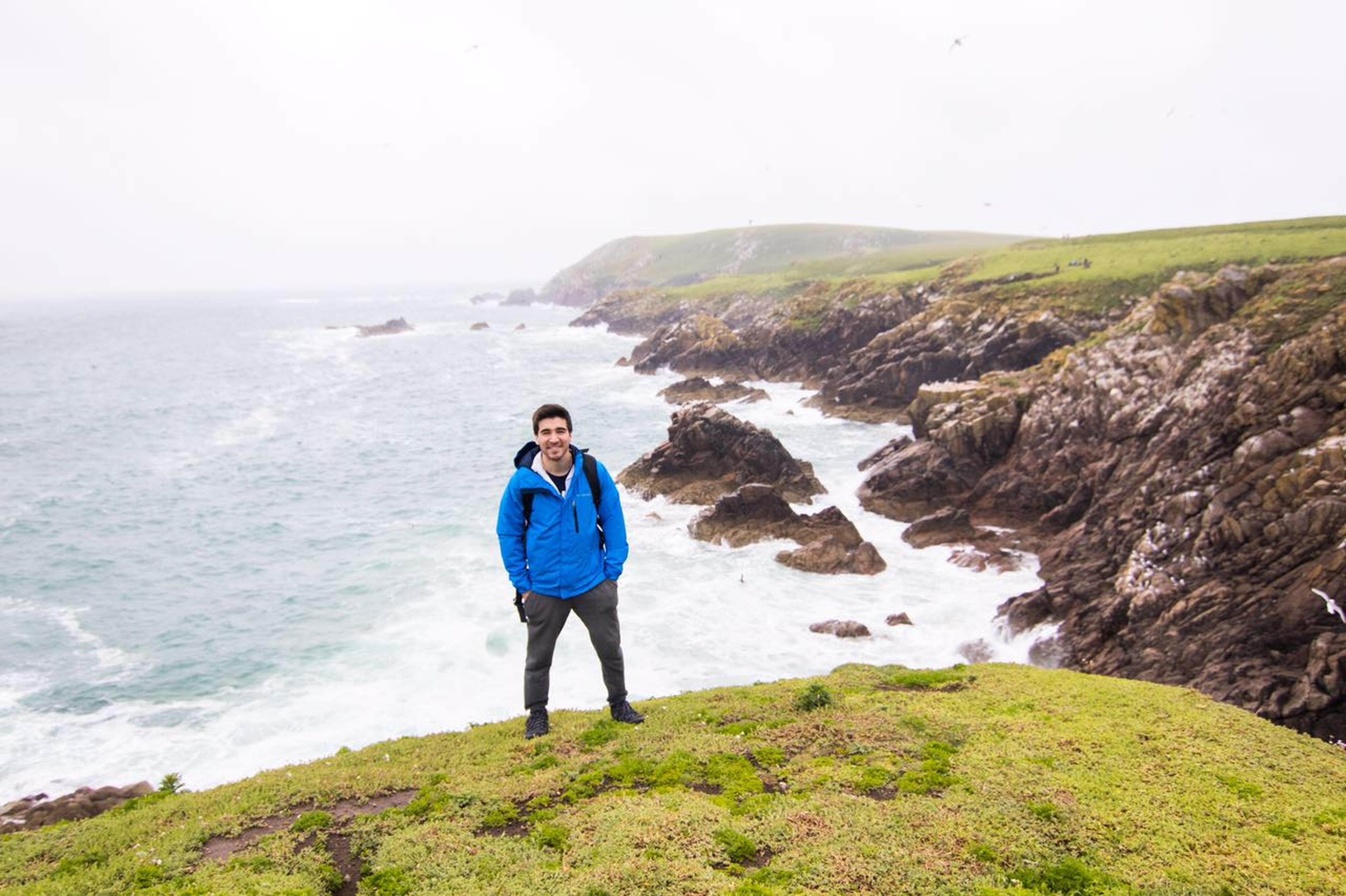 Student standing on green cliffs overlooking the rocky coastline and ocean in Ireland on a cloudy day