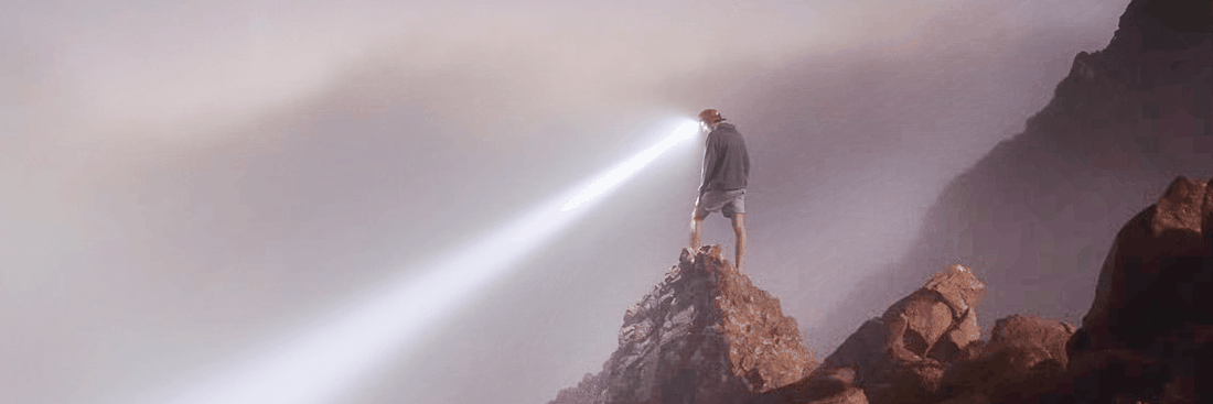 Person steht auf Bergspitze mit Taschenlampe, leuchtet in die Ferne.