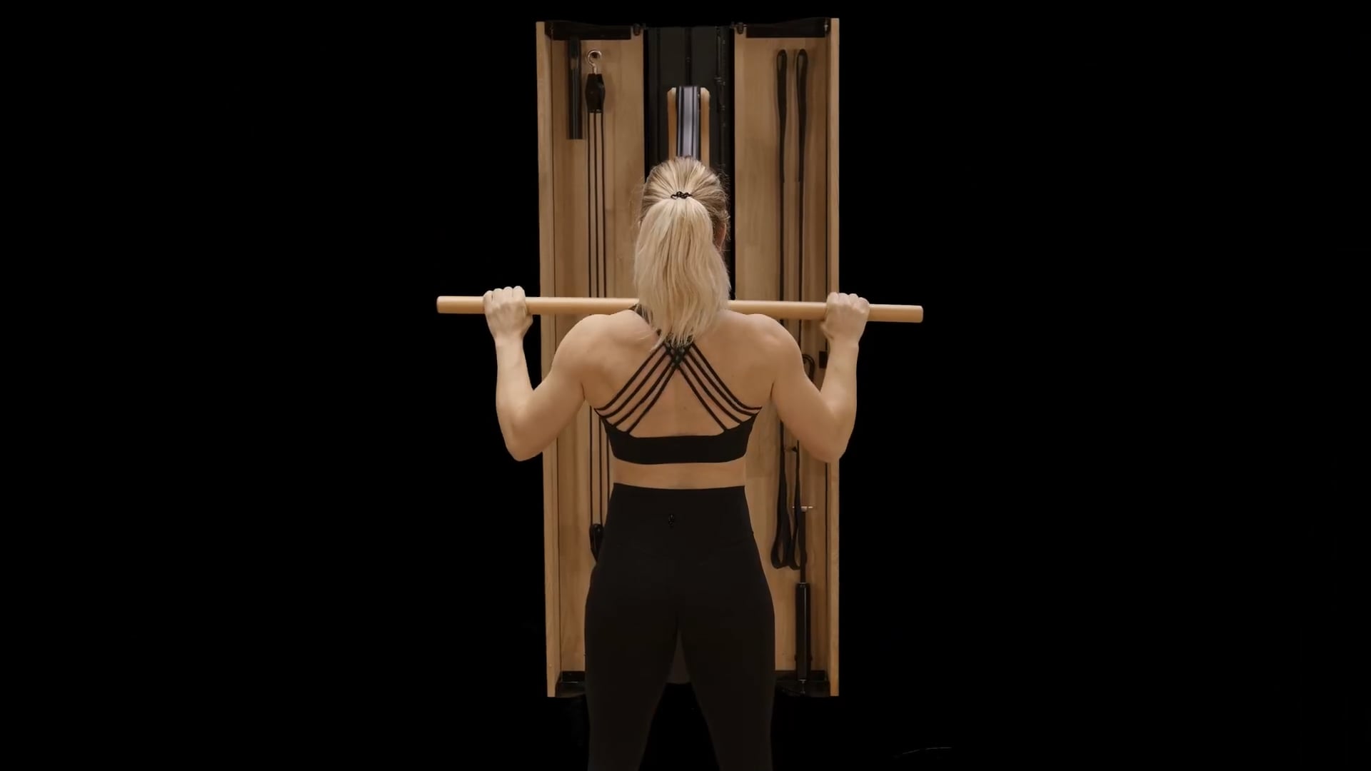 A woman in fitness attire exercises with a wooden resistance machine, holding a bar above her shoulders, viewed from behind against a black background.