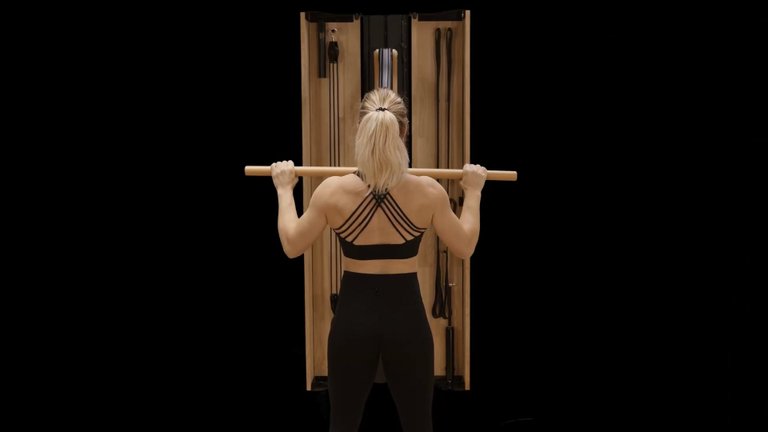 A woman in fitness attire exercises with a wooden resistance machine, holding a bar above her shoulders, viewed from behind against a black background.