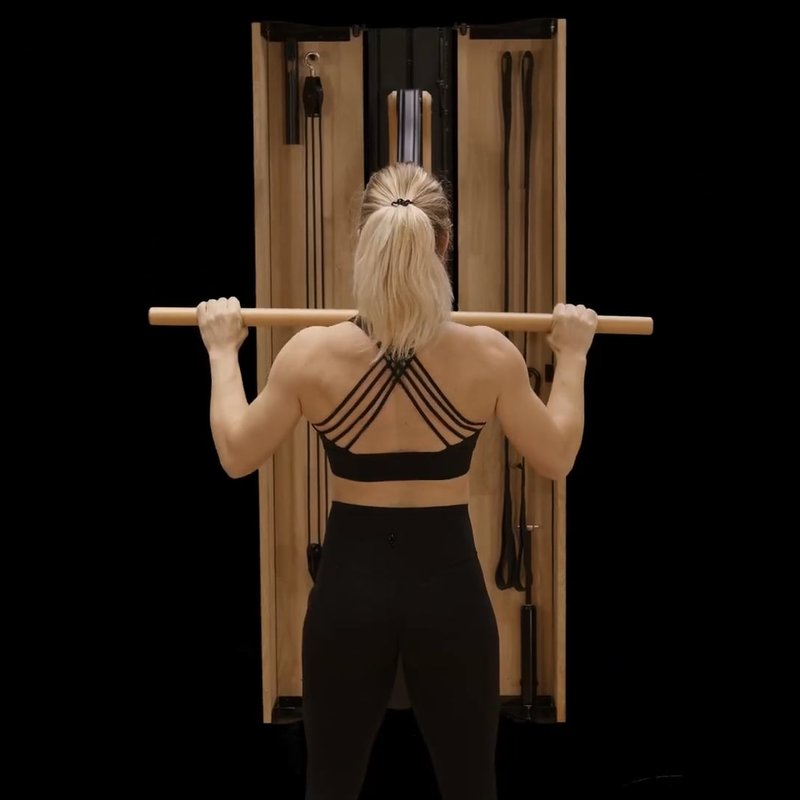 A woman in fitness attire exercises with a wooden resistance machine, holding a bar above her shoulders, viewed from behind against a black background.