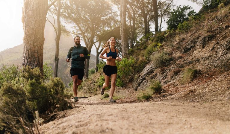 Two people jog on a sunlit dirt trail through a forested area, surrounded by trees and greenery.