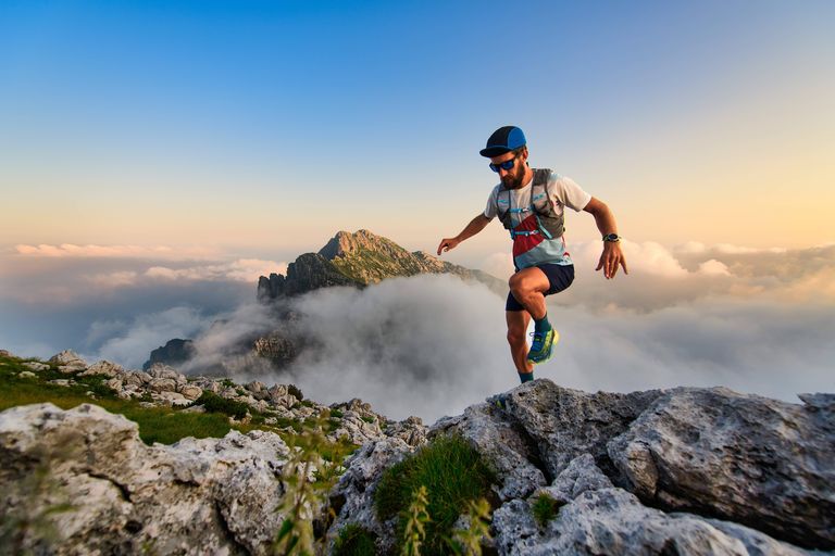 A man in athletic gear and cap runs energetically on rocky terrain, with mountains and clouds in the background under a clear sky.